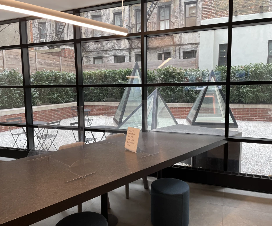 A modern indoor space features a long dark table with clear dividers, facing large windows that offer a view of an outdoor area with glass pyramid structures. The outdoor space includes gravel and green shrubs, with a few bistro tables and chairs. A sign is positioned on the table.