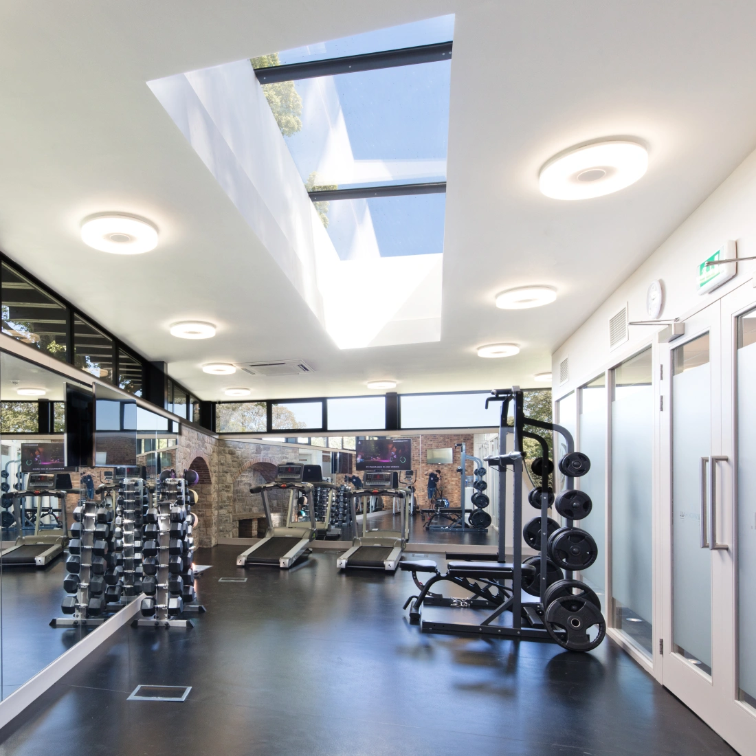 A modern gym interior featuring sleek equipment, large mirrors, and natural light streaming through skylights. Weights are neatly arranged on racks, and fitness machines are lined up against the walls.