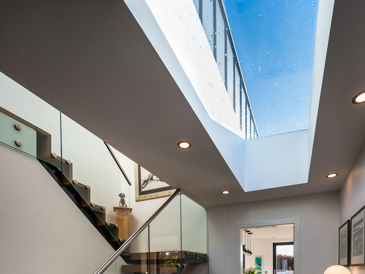 A modern hallway features a striking glass staircase with wooden treads. Natural light pours in through a skylight above. The floor is finished with dark tiles, and a wooden console table holds a vase of yellow flowers, creating a welcoming atmosphere. In the background, a glimpse of an open space with dining furniture is visible.