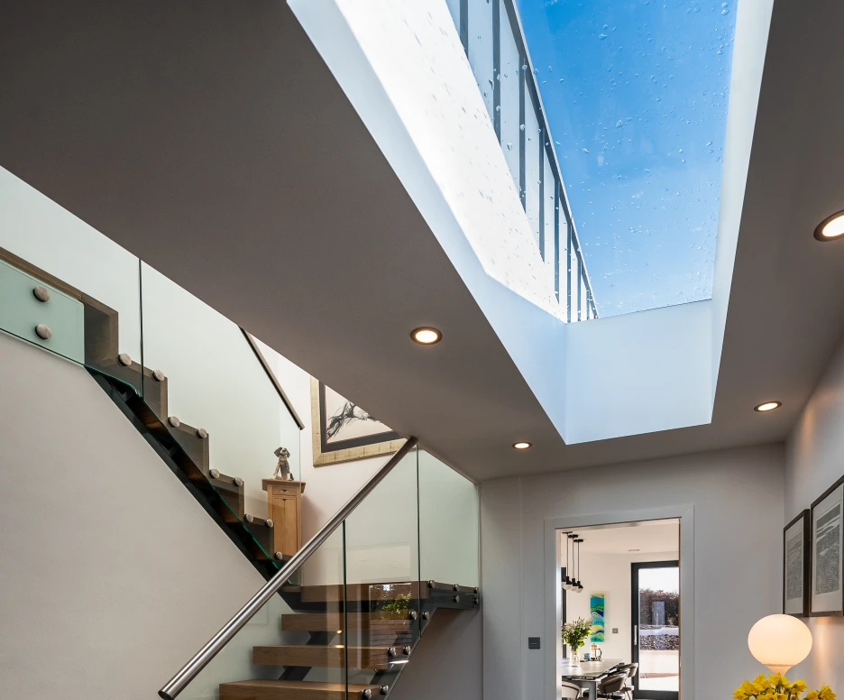 A modern hallway features a striking glass staircase with wooden treads. Natural light pours in through a skylight above. The floor is finished with dark tiles, and a wooden console table holds a vase of yellow flowers, creating a welcoming atmosphere. In the background, a glimpse of an open space with dining furniture is visible.