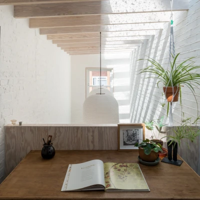 A cozy workspace features a wooden desk with an open book, surrounded by houseplants and framed artwork. Natural light floods in through large windows, illuminating the white brick walls and wooden beams above. A pendant lamp hangs centrally, adding warmth to the space.