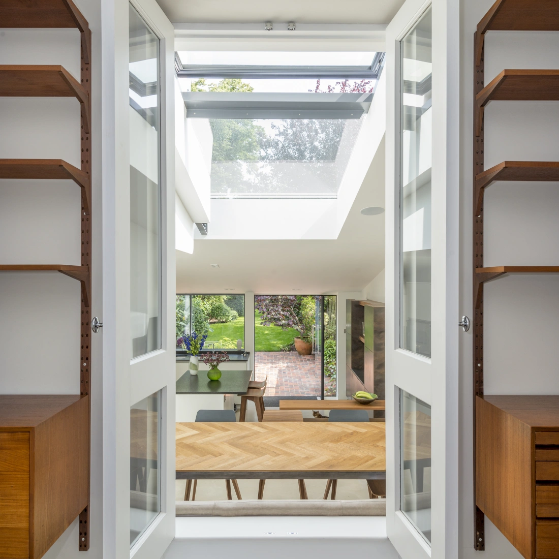 Bright interior view through open double doors, leading to a modern dining area with wooden furniture. Natural light floods in from a skylight above, illuminating the space that opens to a garden outside. Shelves flank the doors, adding to the clean aesthetic.