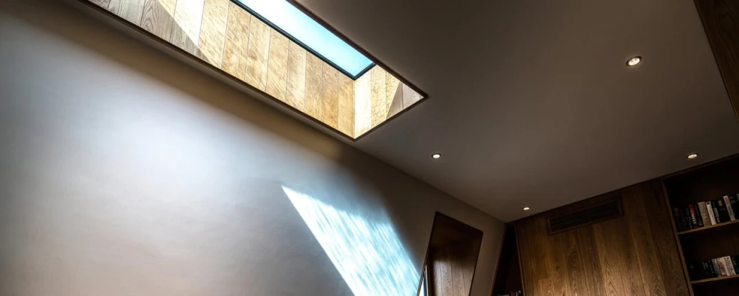 A modern interior featuring a wooden desk and chair, with a large skylight casting geometric light patterns on the wall. Wooden shelving is visible in the background.