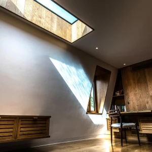 A modern interior featuring a wooden desk and chair, with a large skylight casting geometric light patterns on the wall. Wooden shelving is visible in the background.
