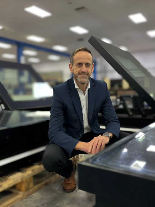 A man in a suit kneels in a warehouse, smiling while surrounded by large glass display cases. The background features shelves and workstations, indicating an industrial setting.