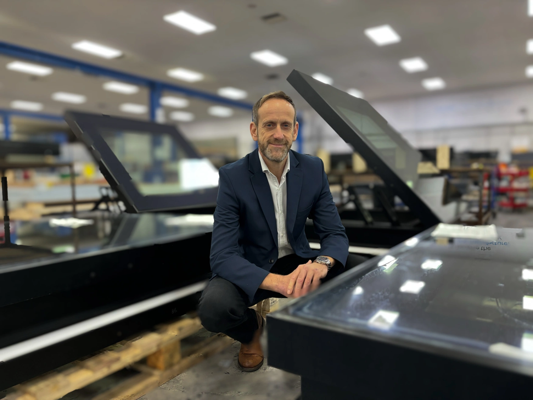 A man in a suit kneels in a warehouse, smiling while surrounded by large glass display cases. The background features shelves and workstations, indicating an industrial setting.