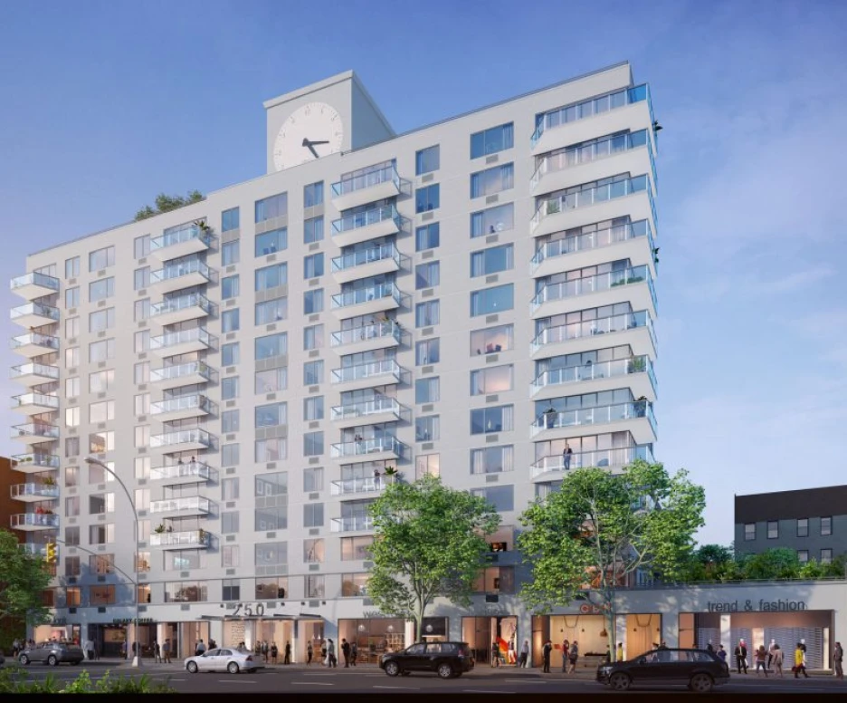 Modern multi-story residential building featuring a large clock on the façade. The building has several balconies and ground-level retail spaces. Trees line the sidewalk, and a clear blue sky provides a backdrop.