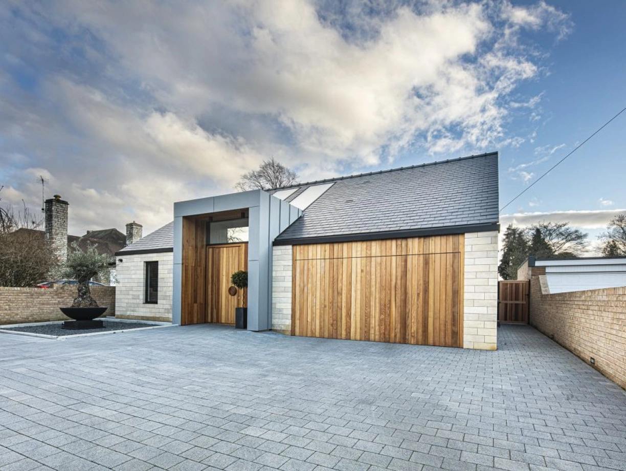 A modern house with a sloped roof, featuring a combination of stone and wood materials. A spacious driveway made of gray pavers leads up to the entrance, which includes a decorative plant near the door. The background shows cloudy skies and a few trees.