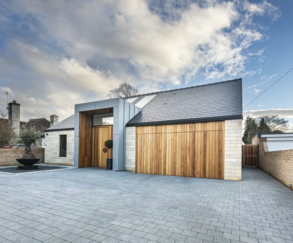 A modern house with a sloped roof, featuring a combination of stone and wood materials. A spacious driveway made of gray pavers leads up to the entrance, which includes a decorative plant near the door. The background shows cloudy skies and a few trees.