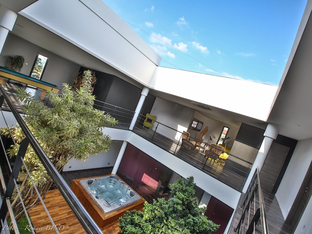 Interiör di un moderni casa con un atrio centrali. Un jacuzzi è visibile in baso, circondati da piante verdi. Mobili eleganti sono disponuti in sale su divers livelli. La luce naturale entra attraverso le finestre grandi.