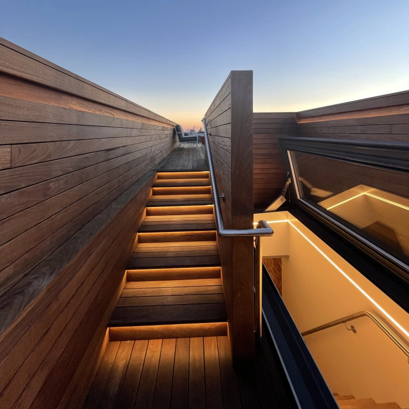 A wooden staircase illuminated by warm lights leads up to a rooftop area at dusk, with the sky transitioning from orange to deep blue.