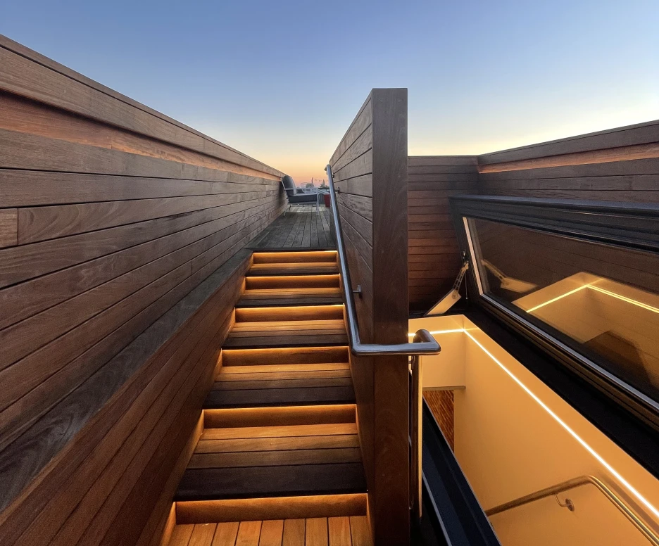 Staircase with illuminated steps leading up to a rooftop, framed by wooden walls, under a clear blue sky at sunset.