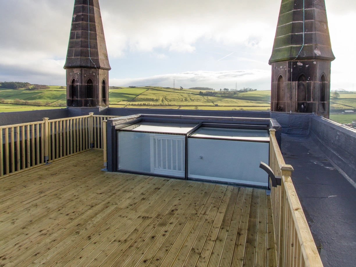 A rooftop view featuring two tall, pointed spires against a cloudy sky. Below, there's a wooden deck and a glass structure, with rolling green hills in the distance.