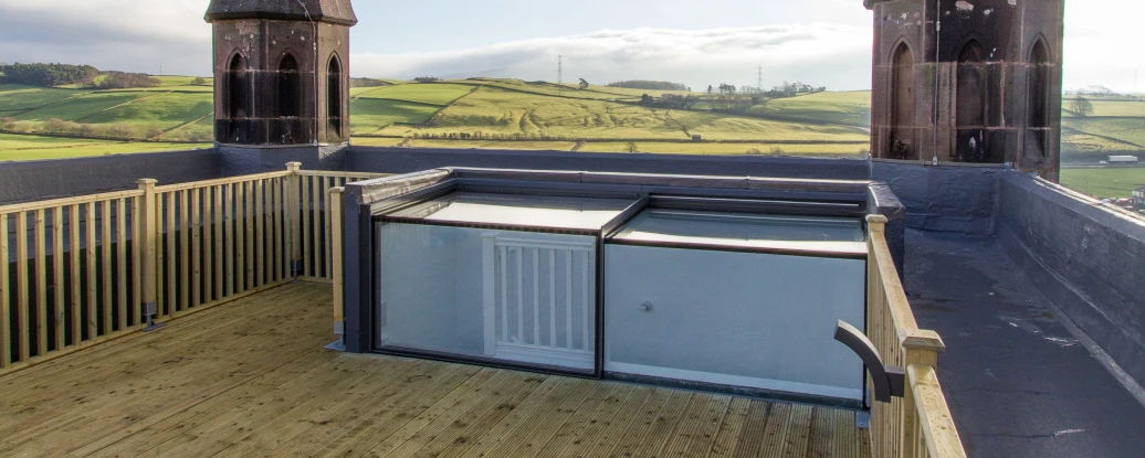 A wooden rooftop deck offering a panoramic view of green hills, featuring two tall, pointed towers with a cloudy sky above.