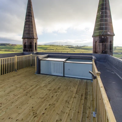 A wooden rooftop deck offering a panoramic view of green hills, featuring two tall, pointed towers with a cloudy sky above.