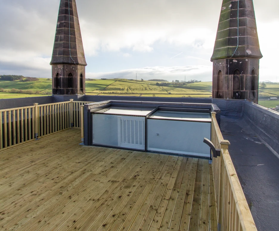 A wooden rooftop deck offering a panoramic view of green hills, featuring two tall, pointed towers with a cloudy sky above.
