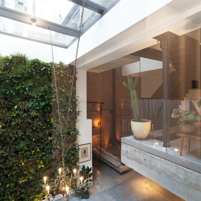 Interior view of a modern space featuring a vertical garden wall, large skylight, and minimalist decor. A staircase is visible in the background alongside stylish furnishings, while a chandelier hangs above a dining table.