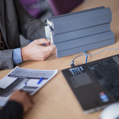 A business professional with a gray suit examines a gray device, while another person holds printed documents beside a laptop on a conference table.