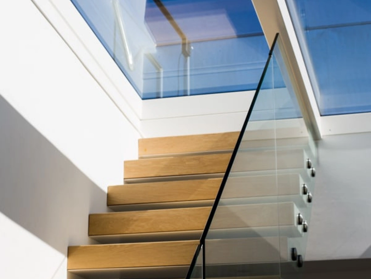 A modern staircase with wooden steps and a glass railing, illuminated by natural light streaming through a skylight above.