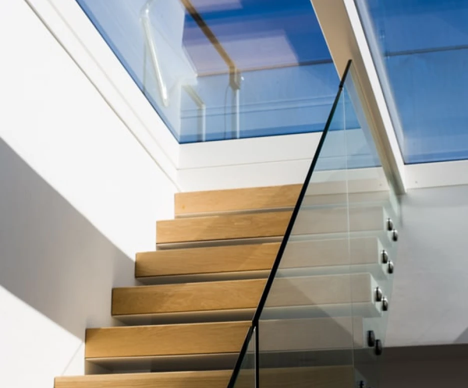 Escalera de madera clara con barandilla de cristal, iluminada por la luz natural que entra por un tragaluz. El fondo muestra un cielo azul despejado.
