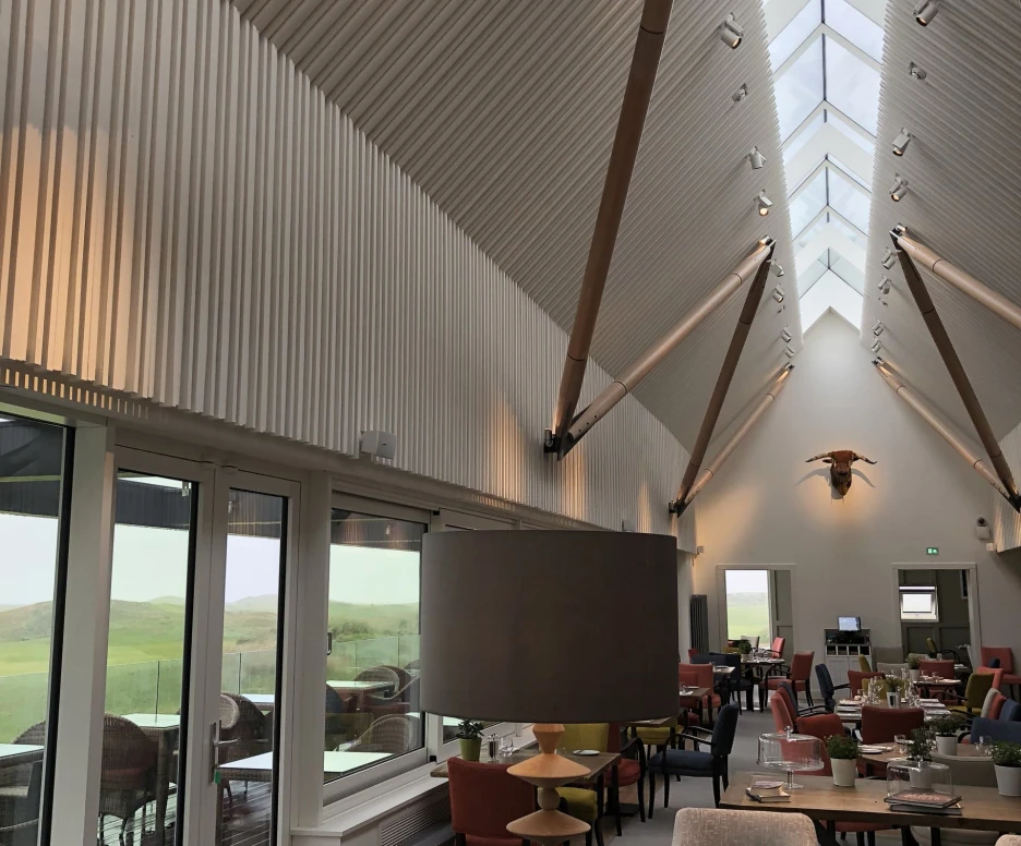 A modern interior of a spacious dining area featuring high ceilings with wooden beams, large windows allowing natural light, and a mix of colorful seating. A stylish lamp stands on a table, and greenery adds a touch of nature to the decor.