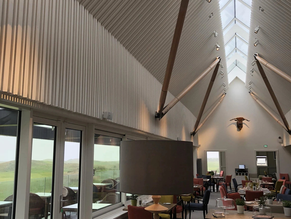 A modern interior of a spacious dining area featuring high ceilings with wooden beams, large windows allowing natural light, and a mix of colorful seating. A stylish lamp stands on a table, and greenery adds a touch of nature to the decor.