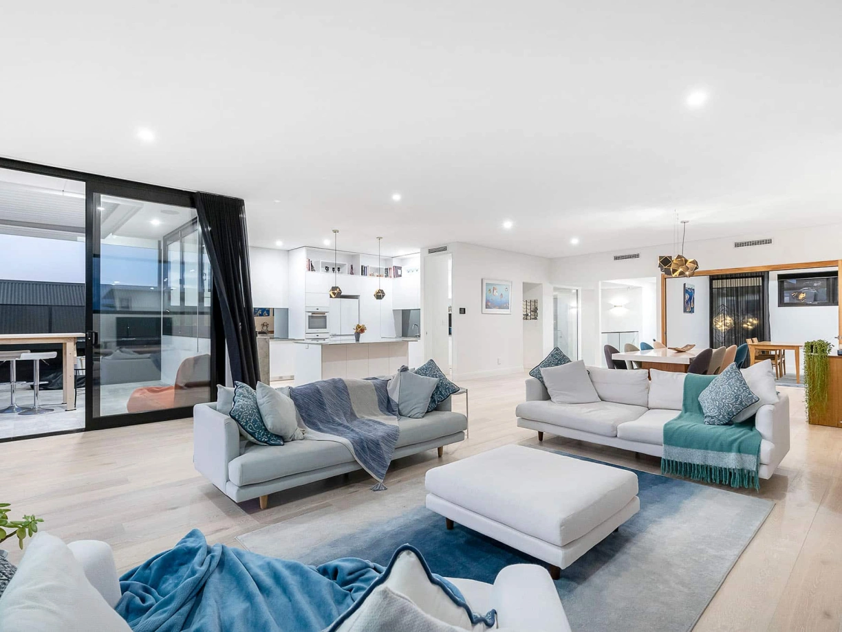 A spacious, modern living room featuring two light-colored couches adorned with decorative pillows and throws. A large ottoman sits in the center, and floor-to-ceiling windows allow natural light to fill the space. In the background, a sleek kitchen is visible, along with a dining area showcasing stylish lighting fixtures.