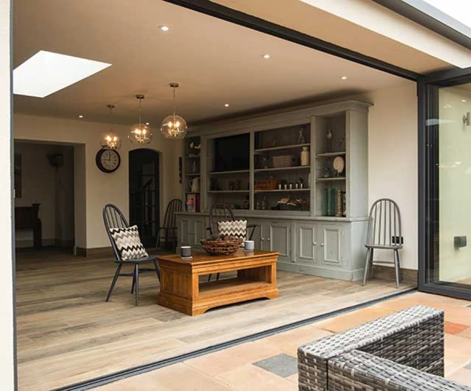 A modern open-concept living space features large glass doors, showcasing a stylish interior with wooden flooring. Two elegant chairs and a rustic wooden coffee table are arranged in front of a gray sideboard filled with decorative items. Soft lighting from pendant lamps adds a warm ambiance.