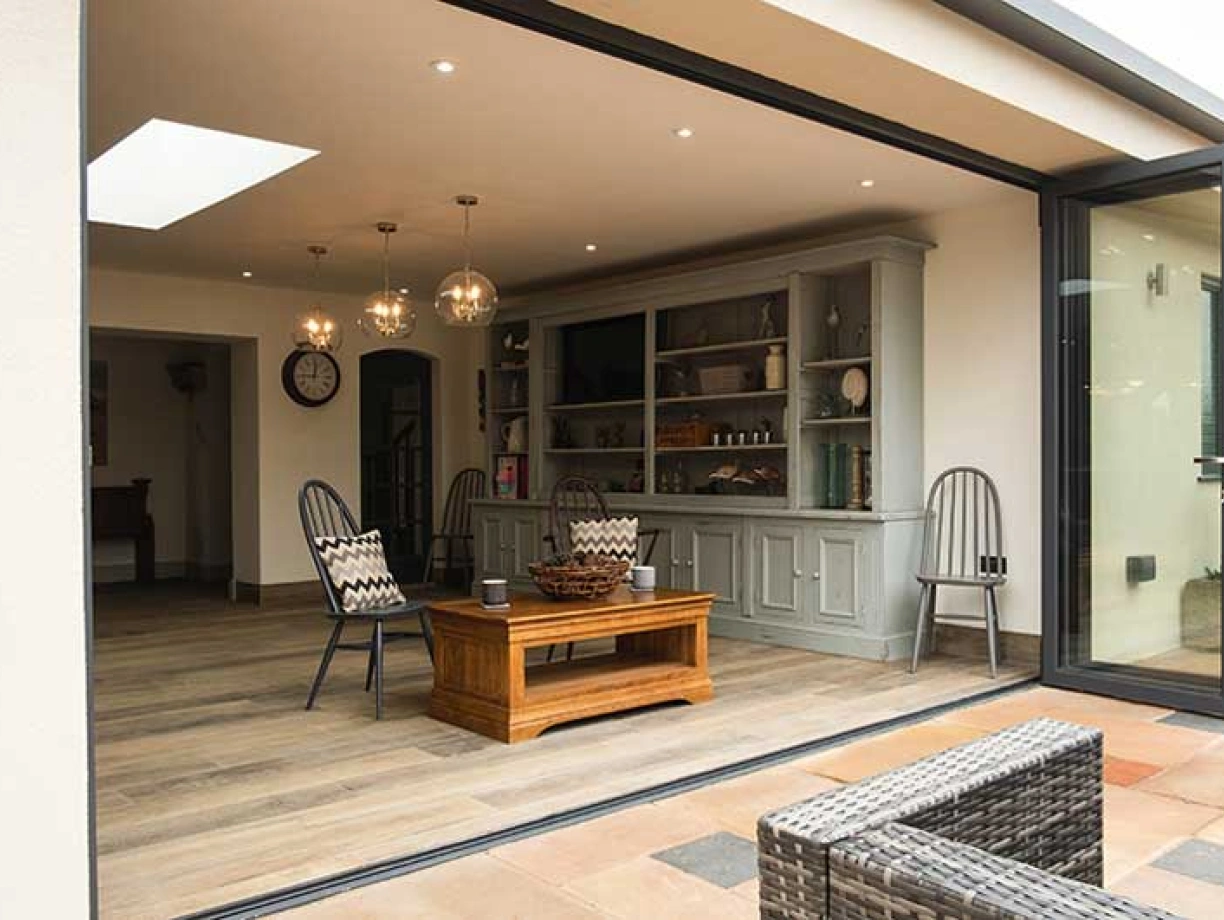 A modern open-concept living space features large glass doors, showcasing a stylish interior with wooden flooring. Two elegant chairs and a rustic wooden coffee table are arranged in front of a gray sideboard filled with decorative items. Soft lighting from pendant lamps adds a warm ambiance.
