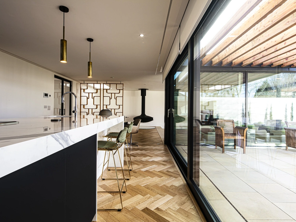 A modern kitchen featuring a large marble island with stylish bar stools, open to a spacious outdoor area. Large glass doors provide a view of a patio with wicker furniture under a wooden pergola, complemented by a cozy fireplace in the background. The flooring showcases a herringbone pattern, enhancing the contemporary aesthetic.