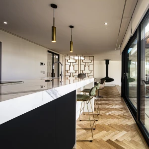 A modern kitchen with a white marble counter and three bar stools, featuring a herringbone wood floor. Large glass windows overlook a patio area with outdoor seating. A stylish, minimalistic design is highlighted by pendant lighting and a sleek fireplace.