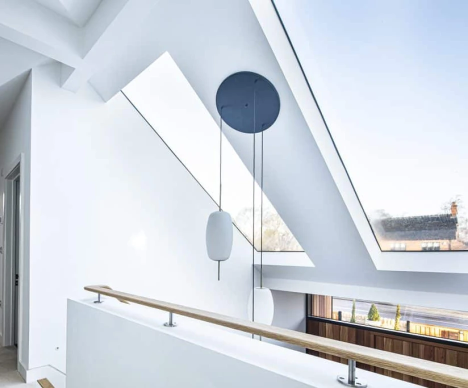 A modern interior view of a loft space featuring slanted ceilings, large skylights, and a wooden railing. Light streams through the windows, illuminating the white walls and minimalistic design. A black circular pendant light hangs from the ceiling.