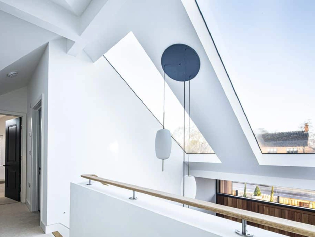 A modern interior view of a loft space featuring slanted ceilings, large skylights, and a wooden railing. Light streams through the windows, illuminating the white walls and minimalistic design. A black circular pendant light hangs from the ceiling.