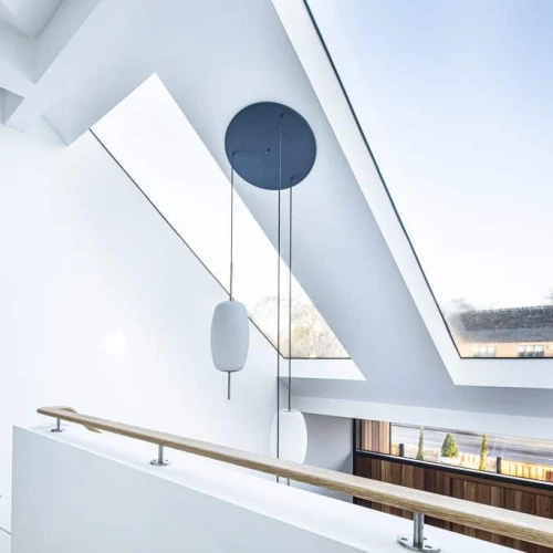A modern interior view of a loft space featuring slanted ceilings, large skylights, and a wooden railing. Light streams through the windows, illuminating the white walls and minimalistic design. A black circular pendant light hangs from the ceiling.