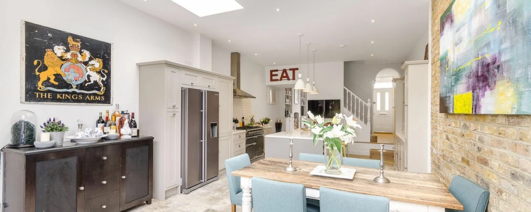A kitchen with a wooden table and blue chairs around it. To the left there is a fridge and cabinets with flowers and bottles on top of it. Above all of this there is 2 fixed skylights