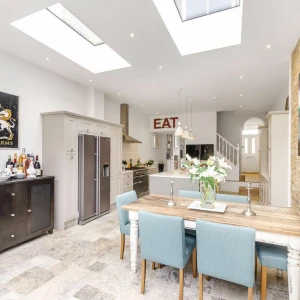 A kitchen with a wooden table and blue chairs around it. To the left there is a fridge and cabinets with flowers and bottles on top of it. Above all of this there is 2 fixed skylights