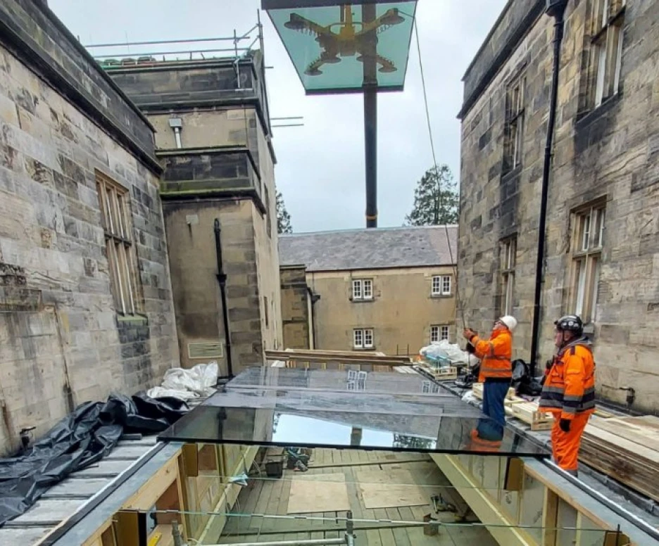Dos trabajadores en ropa de seguridad observan una estructura de vidrio en un espacio en construcción entre dos edificios de piedra. Un gran emblema se encuentra en lo alto, iluminado por la luz natural.