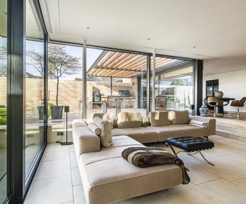 A modern living room with a large sectional sofa, floor-to-ceiling windows, and a view of a patio area. The space features neutral colors, a sleek coffee table, and minimalistic decor. Outdoor seating and a grill are visible through the glass.