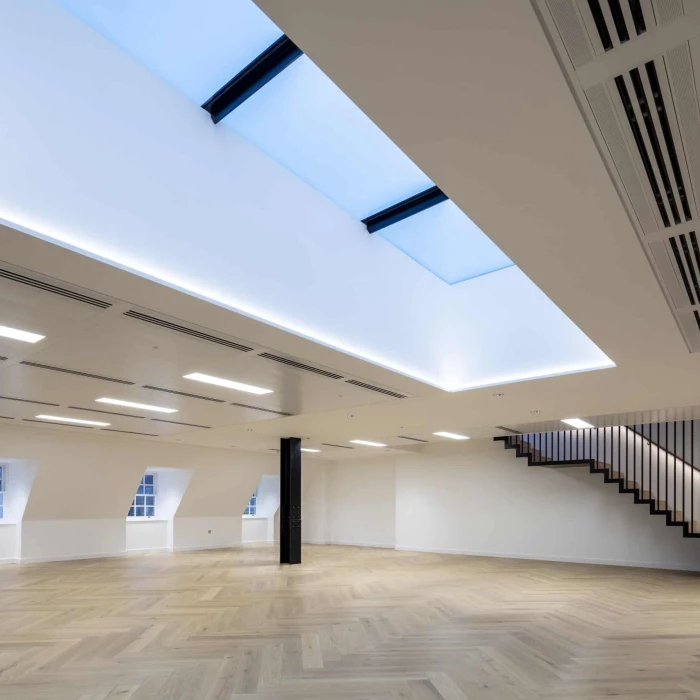 A spacious interior room featuring hardwood flooring and a minimalist design. Skylights illuminate the ceiling, which is adorned with modern lighting fixtures. A wooden staircase with a sleek railing is visible in the corner.