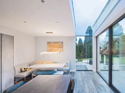 Helle moderne Küche mit einem Essbereich in einer Nische und großen Fenstern, die den Blick auf einen Garten freigeben. Ein minimalistischer Tisch und eine stilvolle Beleuchtung ergänzen das Design.