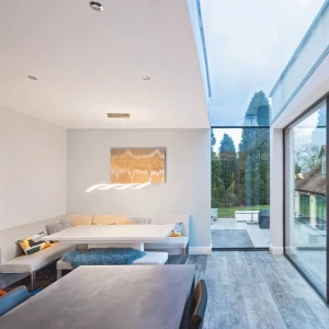 A modern dining area with a minimalist design features a built-in seating nook, a sleek table, and large glass windows showcasing a green outdoor space. Neutral colors dominate the interior, and a stylish light fixture hangs above the table.