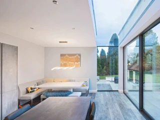 Helle moderne Küche mit einem Essbereich in einer Nische und großen Fenstern, die den Blick auf einen Garten freigeben. Ein minimalistischer Tisch und eine stilvolle Beleuchtung ergänzen das Design.