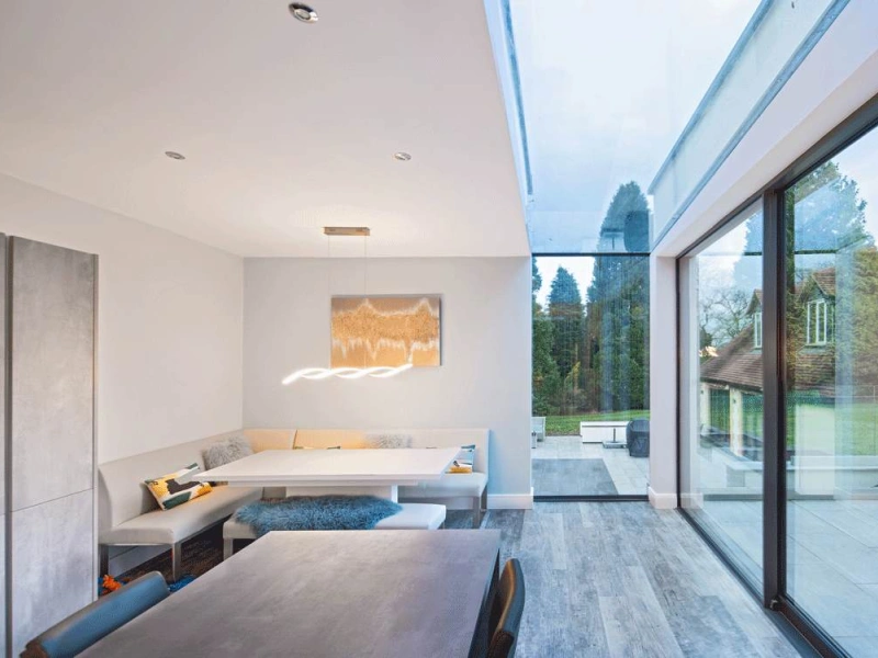 A modern dining area with a minimalist design features a built-in seating nook, a sleek table, and large glass windows showcasing a green outdoor space. Neutral colors dominate the interior, and a stylish light fixture hangs above the table.