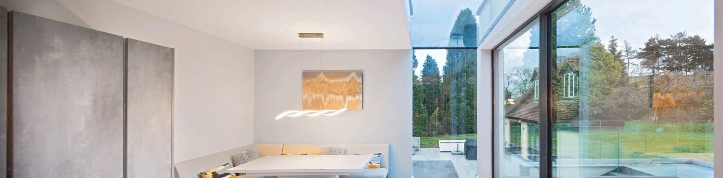 Modern dining area featuring a white booth-style seating, a sleek table, and large glass windows that open to a lush outdoor garden. Natural light floods the space, highlighting the contemporary design and neutral color palette.