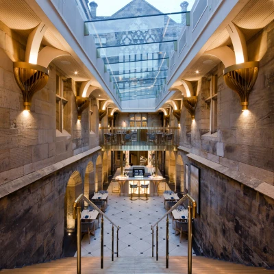 Escalier moderne menant à un espace intérieur lumineux avec des murs en pierre, des luminaires élégants et des tables disposées au centre. Le plafond en verre laisse entrer la lumière naturelle.