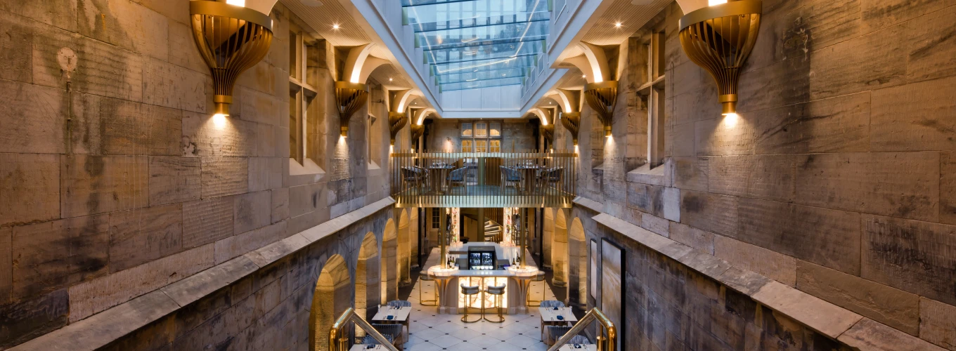 Escalera amplia que desciende a un área con iluminación cálida y paredes de piedra, rodeada de detalles arquitectónicos elegantes. Hay mesas dispuestas en un espacio abierto, con un techo de cristal que permite la entrada de luz natural.