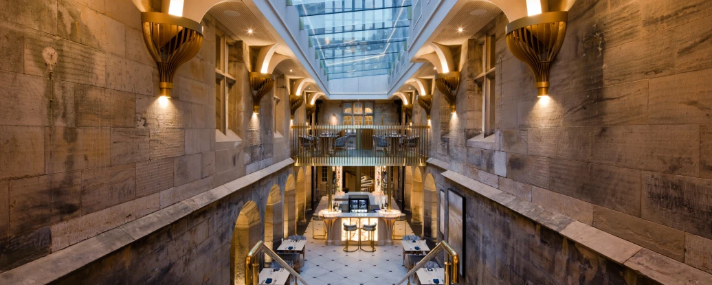 Escalier moderne menant à un espace intérieur lumineux avec des murs en pierre, des luminaires élégants et des tables disposées au centre. Le plafond en verre laisse entrer la lumière naturelle.