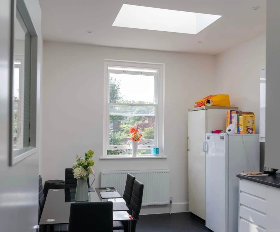 Bright kitchen space with a skylight and a large window. A white refrigerator stands beside a countertop, and a table with black chairs is set up with a vase of flowers. A few food items are on the counter, and sunlight floods the room.