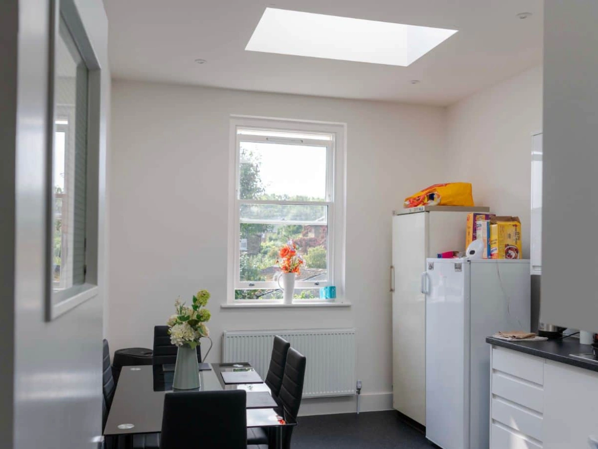 Bright kitchen space with a skylight and a large window. A white refrigerator stands beside a countertop, and a table with black chairs is set up with a vase of flowers. A few food items are on the counter, and sunlight floods the room.