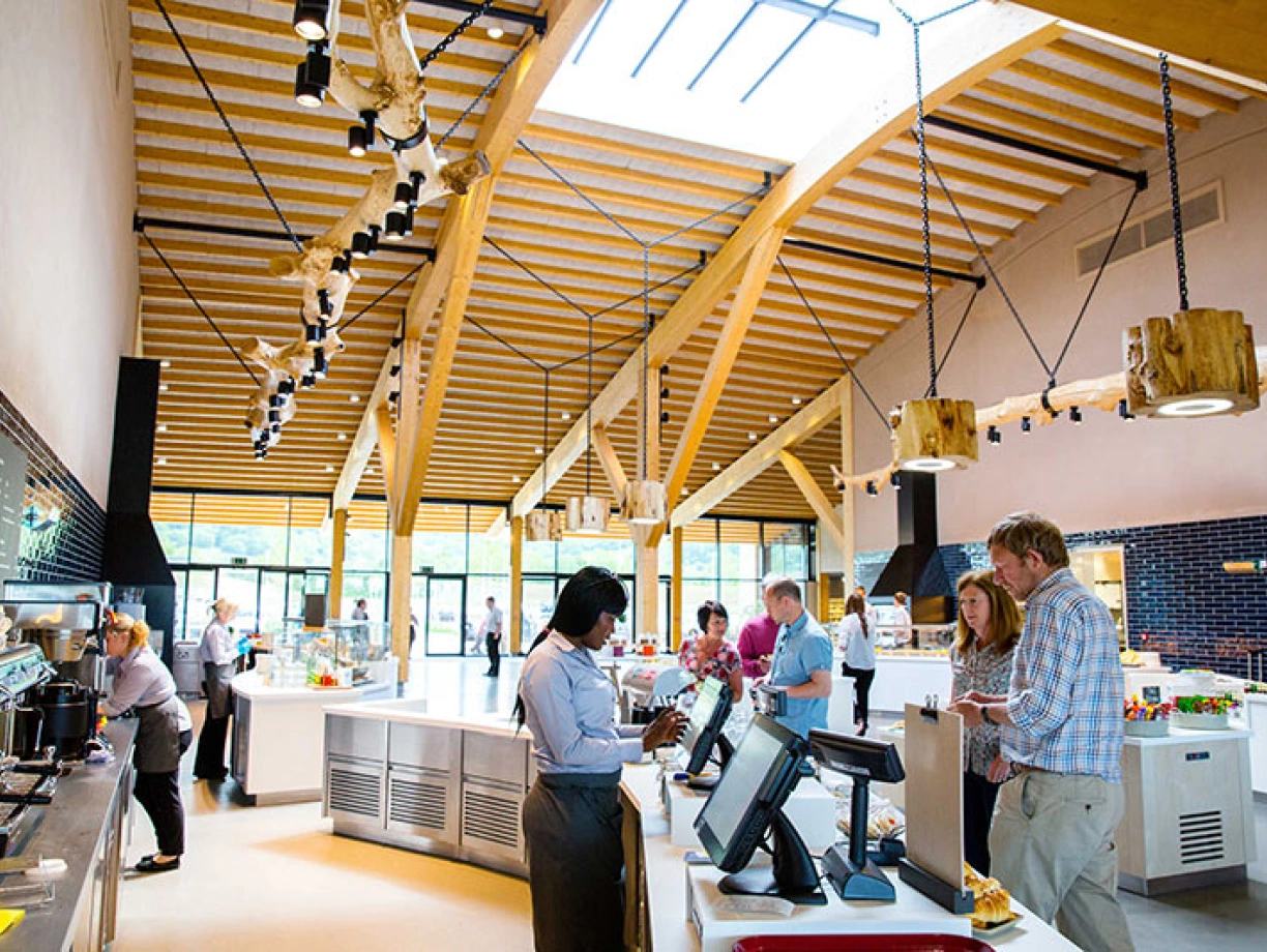 A modern café with a wooden ceiling and large windows. Several people are engaging at the counter, ordering food and drinks. Light fixtures hang from the ceiling, adding a warm ambiance. Various food items are displayed in the background.